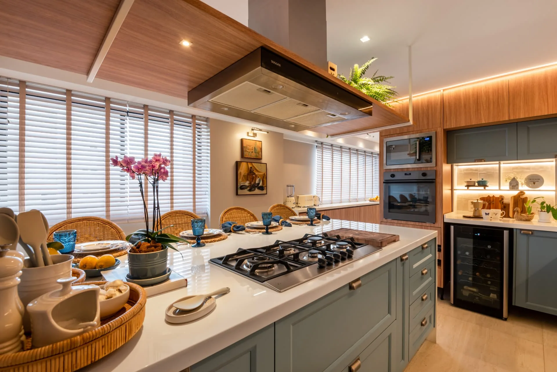 Cozinha Celmar com ilha central em granito, cooktop, armários azul-petróleo e cadeiras de vime